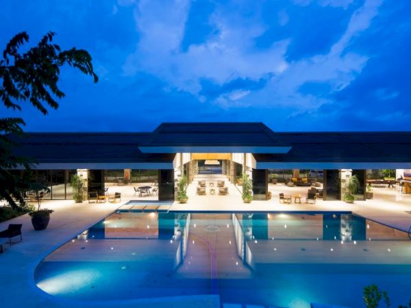 A large, modern building with open spaces is pictured at dusk, featuring a spacious pool in the foreground and seating areas around it.