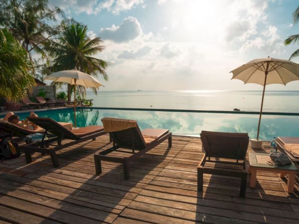 The image shows a serene poolside setting with lounge chairs, umbrellas, palm trees, and a tranquil ocean view under a partly cloudy sky.