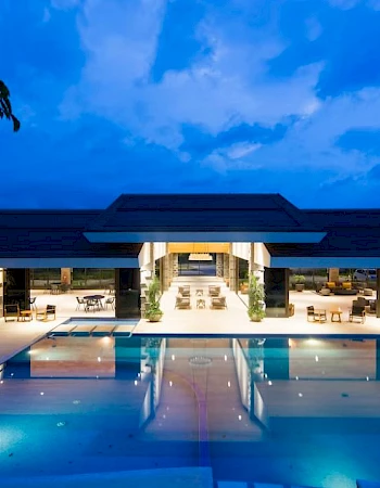 An illuminated outdoor pool area with seating, leading to a modern building under a twilight sky, creating a serene and inviting atmosphere.