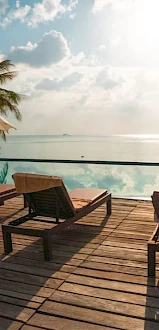 Sunlit deck with lounge chairs, umbrellas, and a serene infinity pool overlooking the ocean. Tropical trees and a partly cloudy sky in the background.