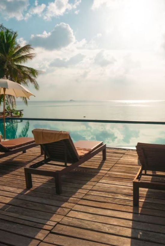 A serene poolside setting with lounge chairs, umbrellas, palm trees, and a calm ocean view under a partly cloudy sky.