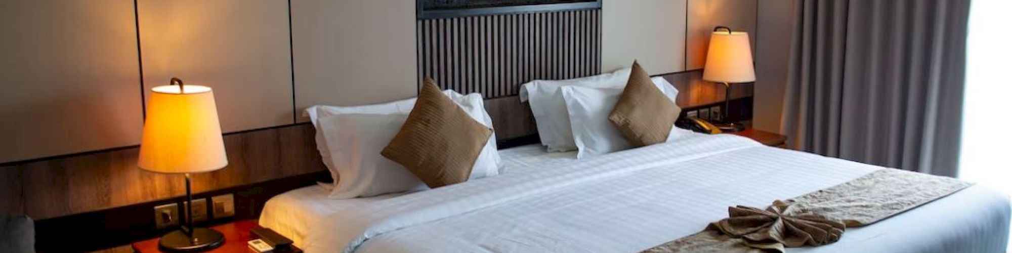 A well-lit hotel room with a large bed, white linens, brown pillows, two bedside tables with lamps, and a fireplace in the background.