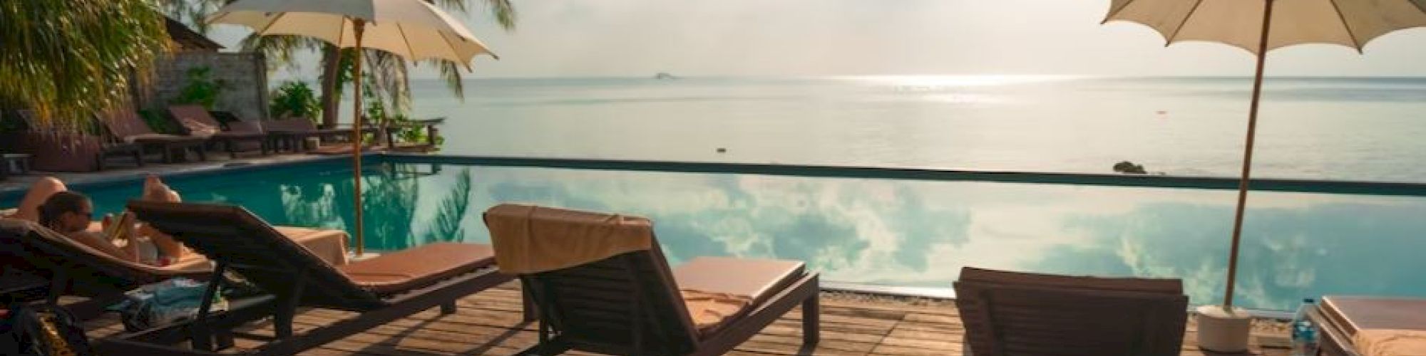A serene poolside scene with lounge chairs, umbrellas, and palm trees, overlooking a calm ocean under a bright sky with scattered clouds.