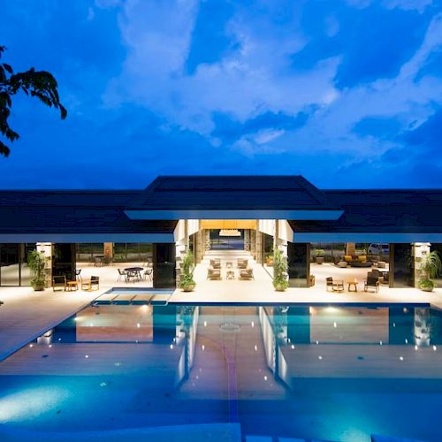 A luxurious, modern building with an illuminated swimming pool and surrounding lounge areas, set against a twilight sky.