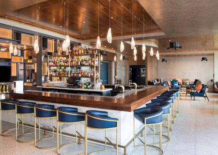 A modern bar with a wooden counter, bar stools, shelves of liquor bottles, hanging lights, and a lounge area with chairs and tables in the background.