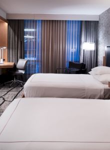 The image shows a modern hotel room with two neatly made beds, a wall-mounted TV, a study desk, and stylish decor with ambient lighting.