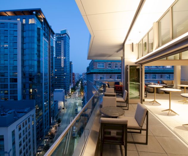 A modern cityscape view from a high-rise balcony at dusk, featuring outdoor seating and large glass buildings lining the streets below.