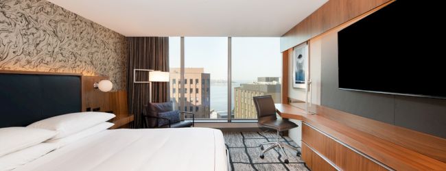 A modern hotel room features a large bed, wall-mounted TV, desk, chair, and panoramic window with an urban view in the background.