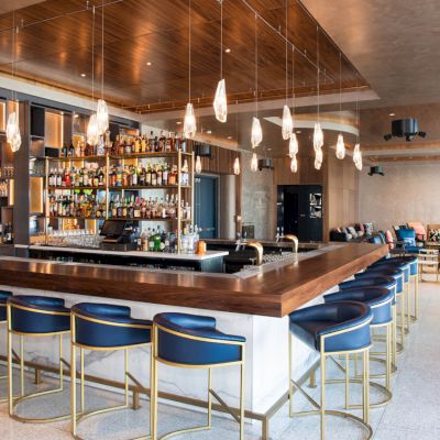 The image shows a modern bar with blue seats, a well-stocked liquor display, pendant lights, and a seating area in the background.