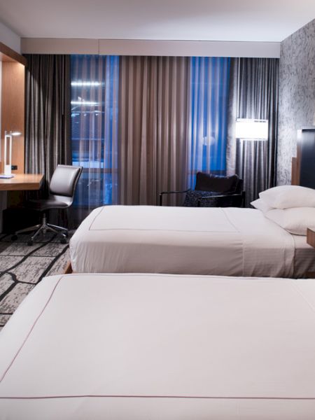The image shows a modern hotel room with two neatly made beds, a desk with a chair, a large TV, and stylish decor with patterned walls and carpeting.
