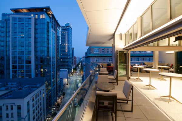A modern balcony at dusk with city views, featuring seating and tables. The city's buildings are illuminated, adding to the urban atmosphere.
