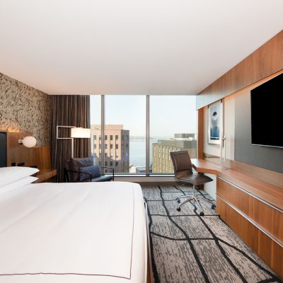A modern hotel room with a large bed, a desk with a chair, a flat-screen TV on the wall, and a window with a cityscape view.