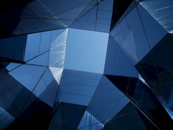 The image shows an upward view of modern, mirrored, geometric architectural structures converging towards the sky, creating a striking design.