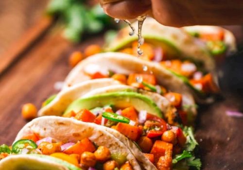 The image shows a hand squeezing lime juice over a row of vibrant tacos filled with vegetables and chickpeas, placed on a wooden surface.