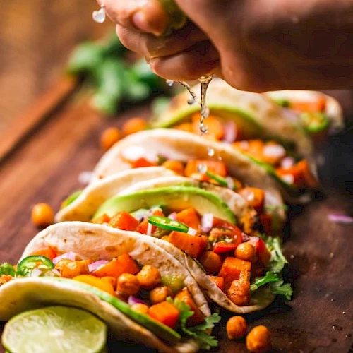 The image shows a hand squeezing lime juice over a row of vibrant tacos filled with vegetables and chickpeas, placed on a wooden surface.