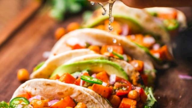 The image shows a hand squeezing lime juice over a row of vibrant tacos filled with vegetables and chickpeas, placed on a wooden surface.