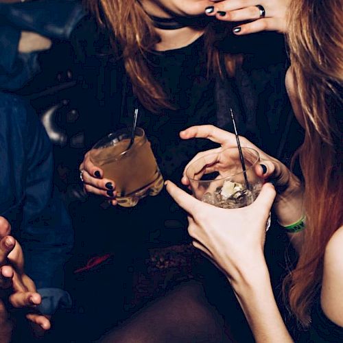This image shows people socializing, holding drinks. They appear to be having a lively conversation in a dimly lit setting.