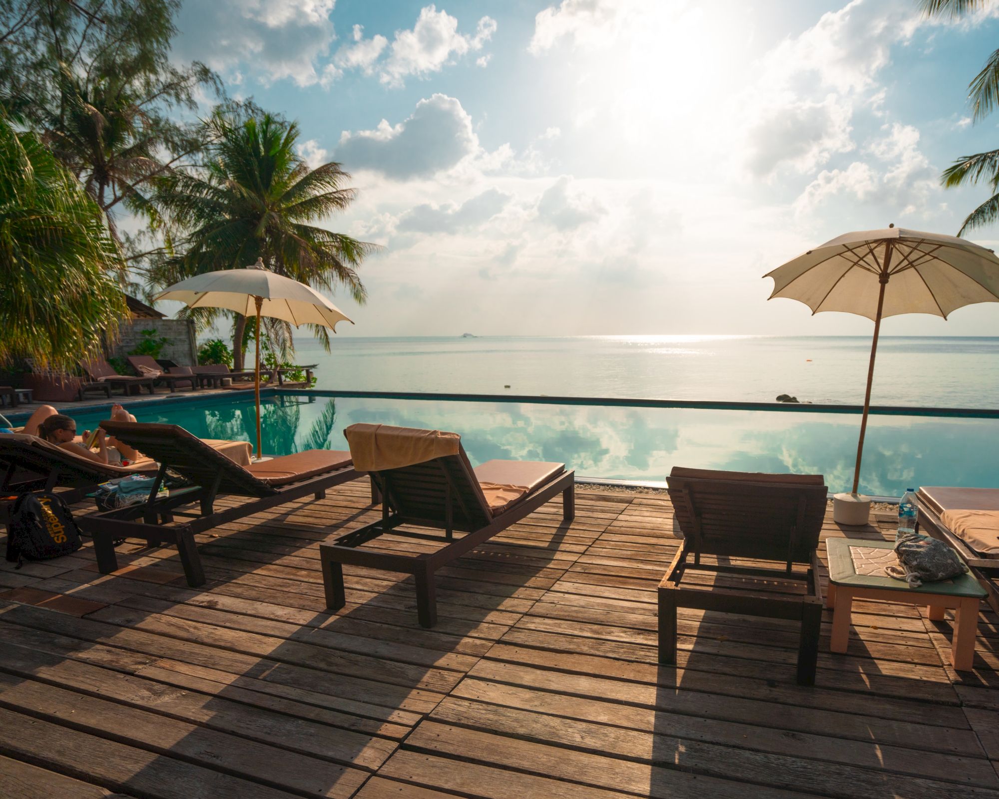 A serene beachside scene with sun loungers, umbrellas, a wooden deck, palm trees, and a calm sea under a partly cloudy sky at sunset.