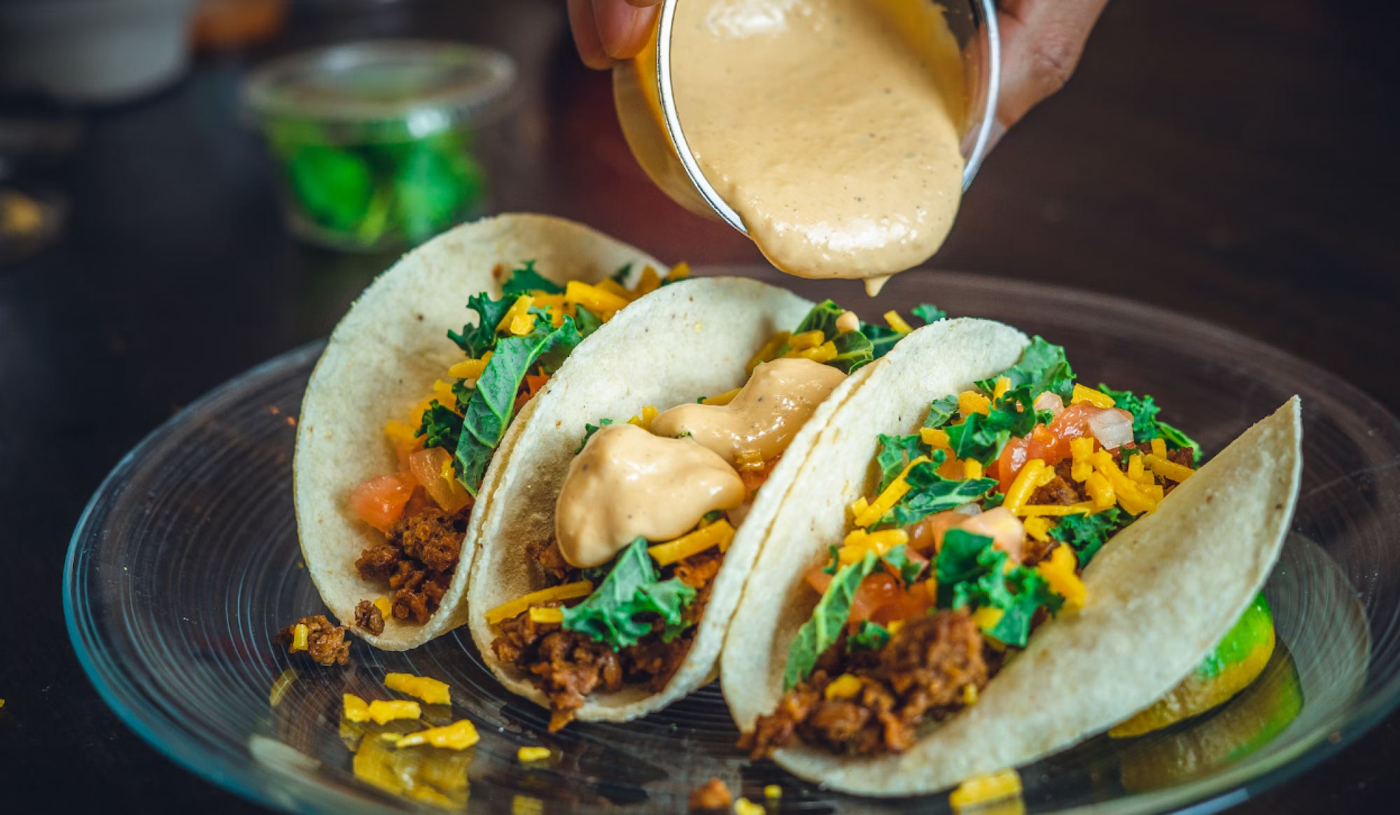 Three tacos on a plate filled with ground meat, cheese, lettuce, and tomatoes, with sauce being poured over them.