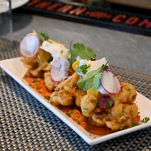 A rectangular white plate with a gourmet dish, featuring what appears to be fried cauliflower, garnished with radishes, greens, and a red sauce.