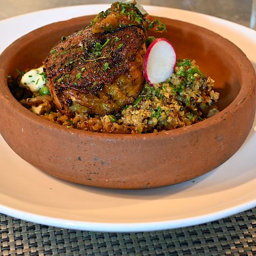 This image shows a dish in a brown bowl with what appears to be seasoned meat on top of a bed of grains, garnished with herbs and a radish slice.