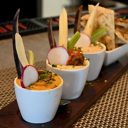 The image shows a serving board with three small cups of dip garnished with vegetables and a dish of assorted thinly sliced bread or crackers.