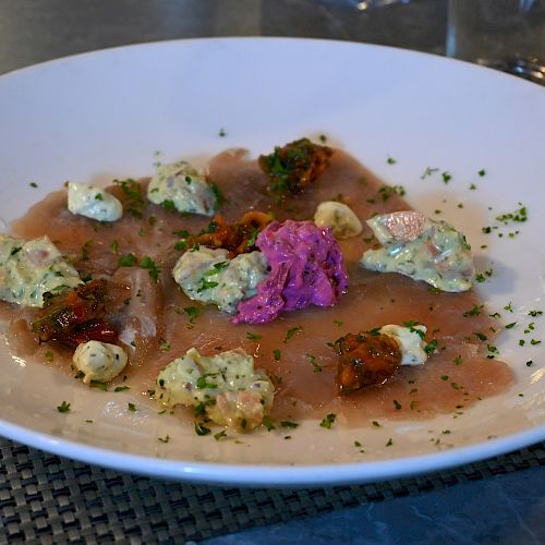 A gourmet dish on a white plate featuring thin slices of meat, colorful dollops of sauces, garnished with herbs, and served on a woven placemat.