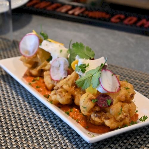 The image shows a rectangular white plate with a gourmet dish, featuring fried cauliflower topped with fresh herbs, radish slices, and sauce.