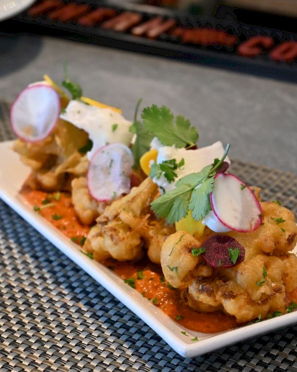 The image shows a rectangular white plate with a gourmet dish, featuring fried cauliflower topped with fresh herbs, radish slices, and sauce.