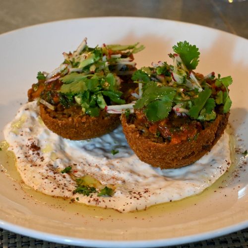 The image shows a plated dish of two falafel patties topped with herbs and served on creamy sauce in a white bowl, garnished with fresh cilantro.