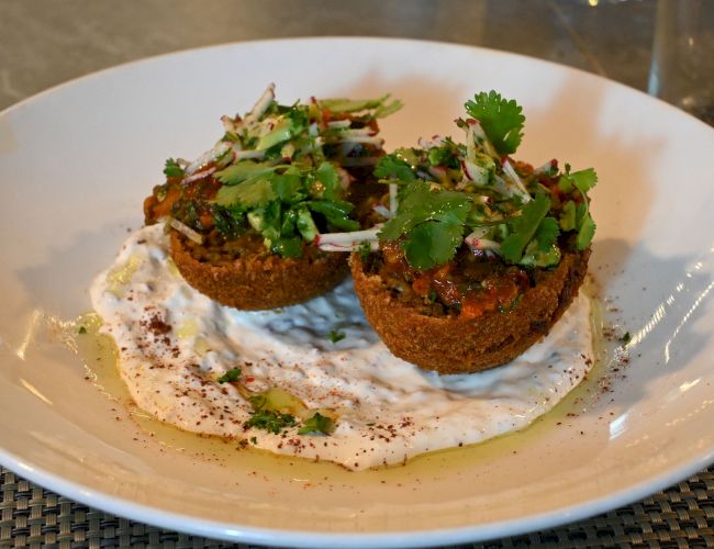 The image shows a plated dish of two falafel patties topped with herbs and served on creamy sauce in a white bowl, garnished with fresh cilantro.