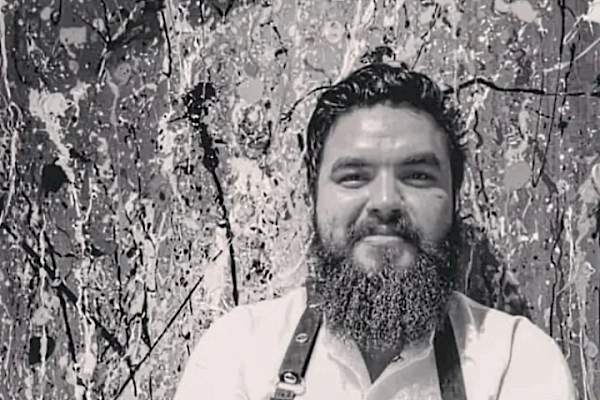 A bearded man with crossed arms is wearing a white shirt and apron, standing in front of a lively, abstract splatter-painted background.