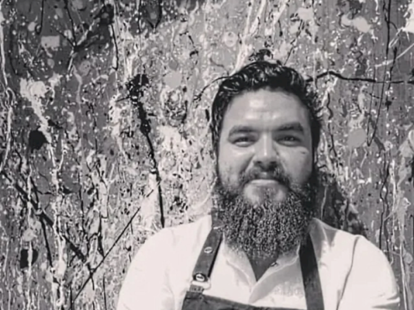 A bearded man with crossed arms is wearing a white shirt and apron, standing in front of a lively, abstract splatter-painted background.