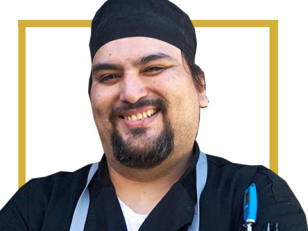 A smiling man is wearing a black chef's hat and jacket. He has a pen in his pocket and is standing against a simple background.