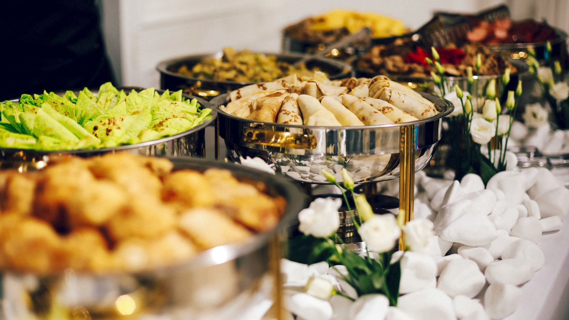 The image shows a buffet setup with various dishes, including spring rolls, leafy greens, and other foods, presented in metal chafing dishes.