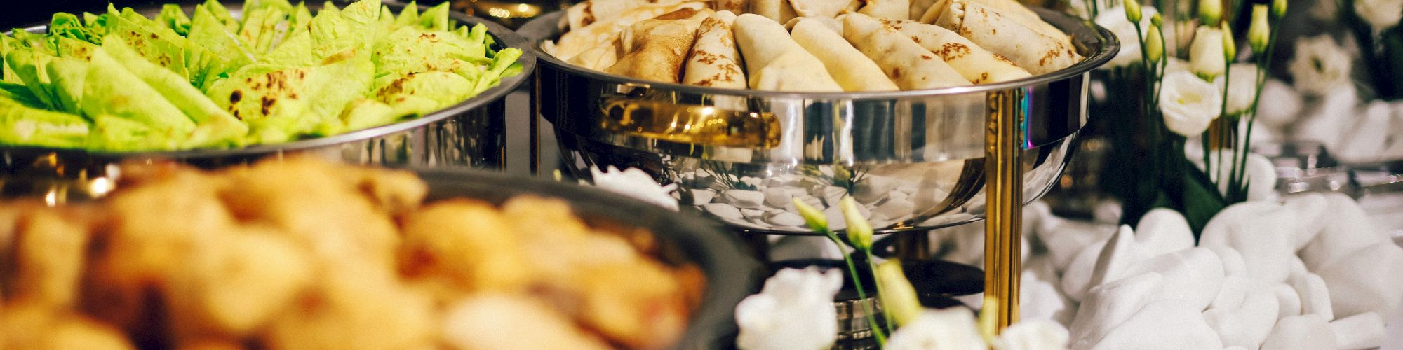 The image shows a buffet setup with various dishes, including spring rolls, leafy greens, and other foods, presented in metal chafing dishes.