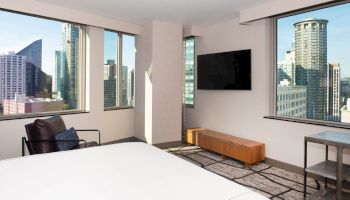 A modern hotel room with large windows, city view, TV, chair, bed, and a side table, showcasing urban skyscrapers outside the windows.