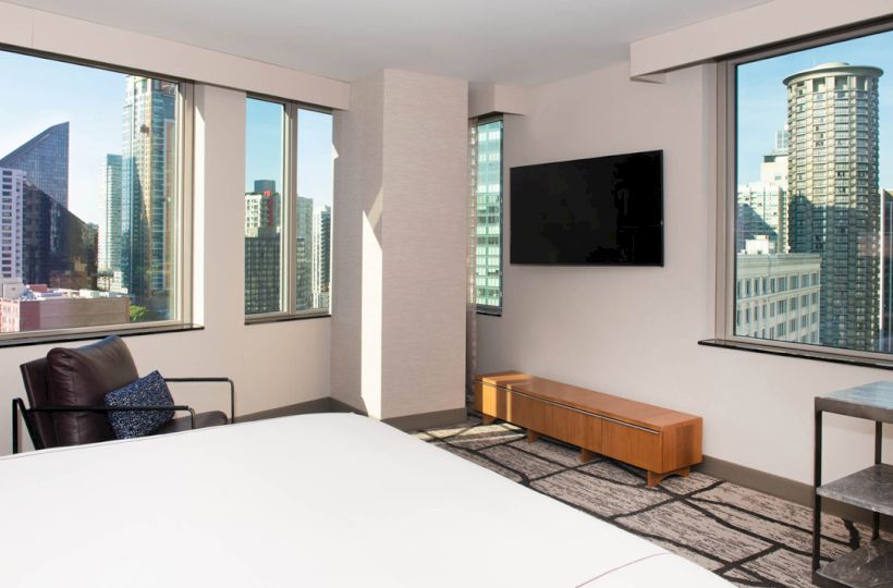 A modern hotel room with large windows, city view, TV, chair, bed, and a side table, showcasing urban skyscrapers outside the windows.