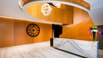 A modern lobby area with a curved wooden ceiling, a large clock on the wall, a marble reception desk, and a vase of flowers.
