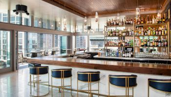 A modern bar with a wooden counter, blue barstools, a wide variety of liquor bottles on shelves, and large windows providing a city view ending the sentence.
