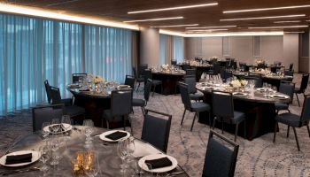 A formal dining room with round tables set for an event, featuring elegant table settings and blue cushioned chairs, under soft ambient lighting.