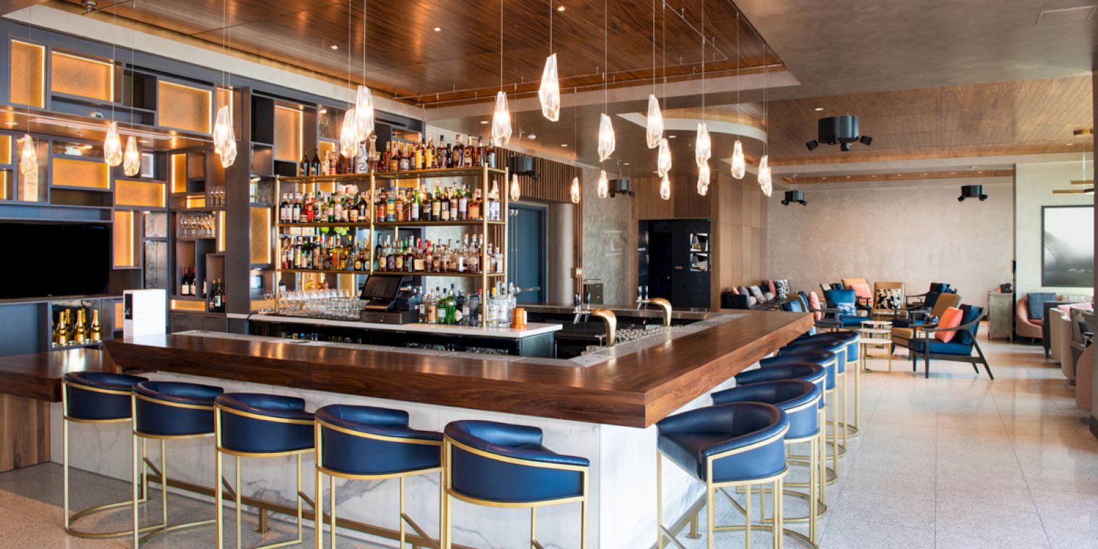 A modern bar with a large selection of bottles, blue stools, sleek lighting, and a lounge area in the background.
