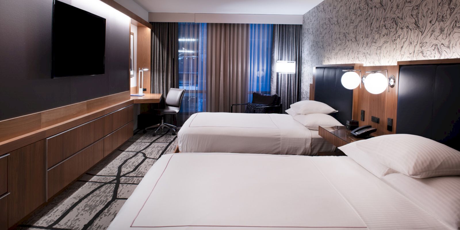 The image shows a modern hotel room with two neatly made beds, a desk with a chair, a wall-mounted TV, and contemporary decor.