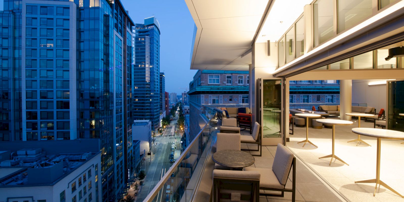An outdoor patio with tables and chairs next to a city view at dusk. Tall buildings line both sides of the street, lit up for the evening.
