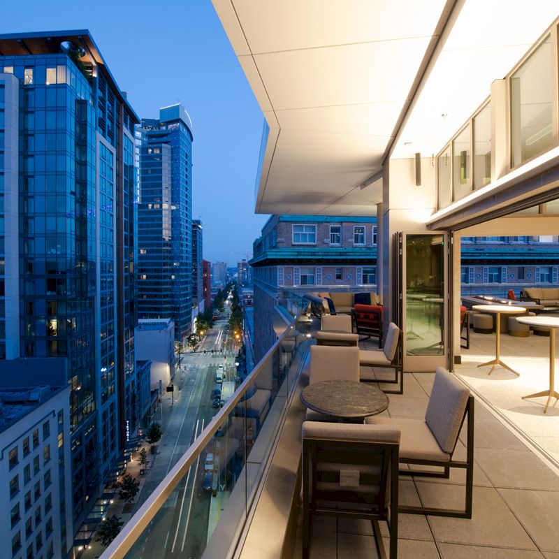 A modern balcony with tables and chairs overlooks an urban cityscape with tall buildings and evening lights, creating a tranquil ambiance.