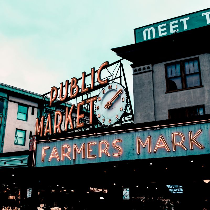 A neon sign for a public farmers market located on a building, with a retro-style clock. The surrounding area includes other buildings and shops.