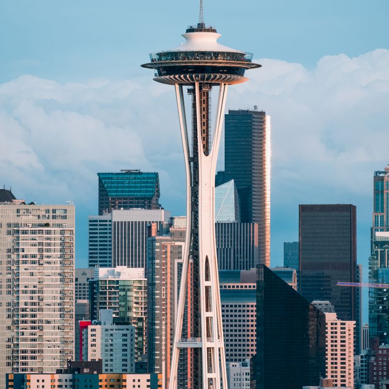 The image shows the Space Needle with a backdrop of modern skyscrapers under a partly cloudy sky in Seattle, Washington.