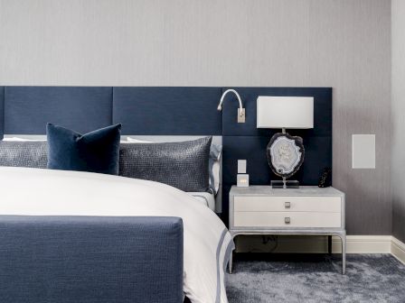 A modern bedroom features a blue upholstered bed, white bedding, a bedside table with a lamp, decorative items, and a cozy gray carpet.