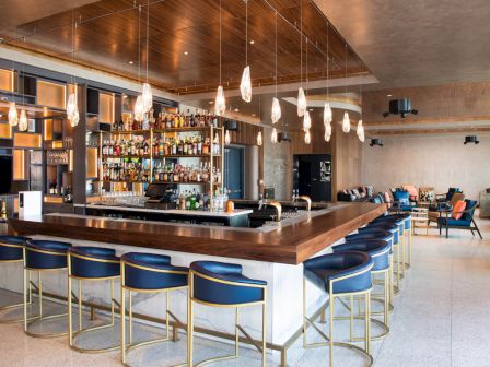 A modern bar area with a wooden counter, blue cushioned stools, pendant lights, and a fully stocked bar, surrounded by a stylish lounge seating area.