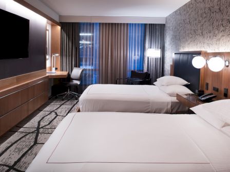 A modern hotel room with two beds, a TV on the wall, a desk area, lamps, and large windows with curtains.